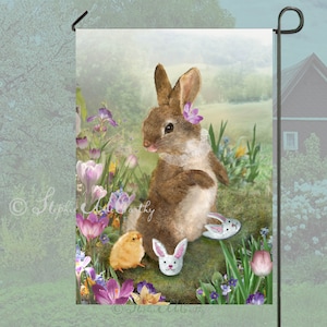May include: A brown bunny rabbit with a purple flower in its ear sits on a rock with a yellow chick and a white bunny slipper. The background is a field of purple and yellow flowers.