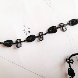 May include: Black braided trim with a repeating pattern of small oval shapes and bows. The trim is on a white surface.