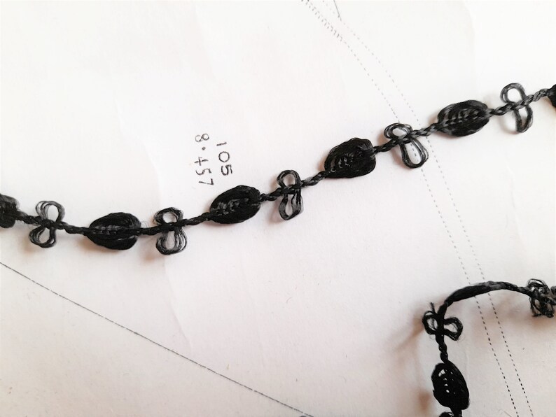 May include: Black braided trim with a repeating pattern of small oval shapes and bows. The trim is on a white surface.