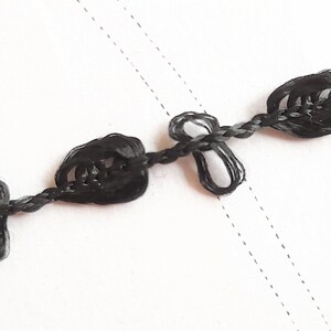May include: Black braided trim with decorative knots. The trim is made of a thin, black cord with small, knotted loops spaced evenly along its length.