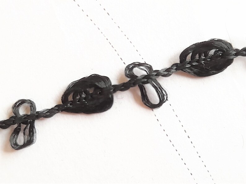 May include: Black braided trim with decorative knots. The trim is made of a thin, black cord with small, knotted loops spaced evenly along its length.