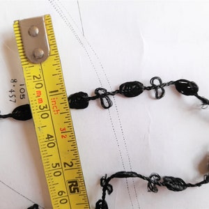 May include: Black trim with a knotted design, measured with a yellow tape measure marked in inches and millimeters. The trim is approximately 3 inches long.