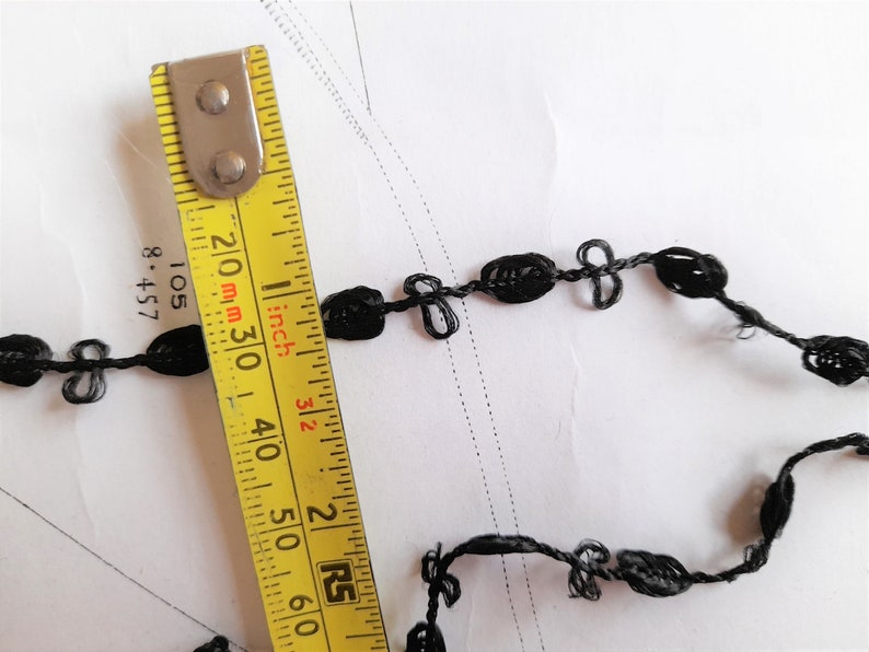 May include: Black trim with a knotted design, measured with a yellow tape measure marked in inches and millimeters. The trim is approximately 3 inches long.