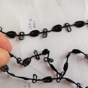 May include: Black trim with a knotted design. The trim is made of a thin, flexible material and has a series of small, evenly spaced knots.