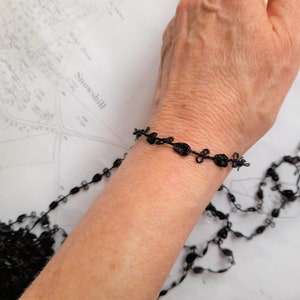 May include: A black lace bracelet with a delicate design. The bracelet is made of thin, black thread that is woven into a series of small, intricate knots.