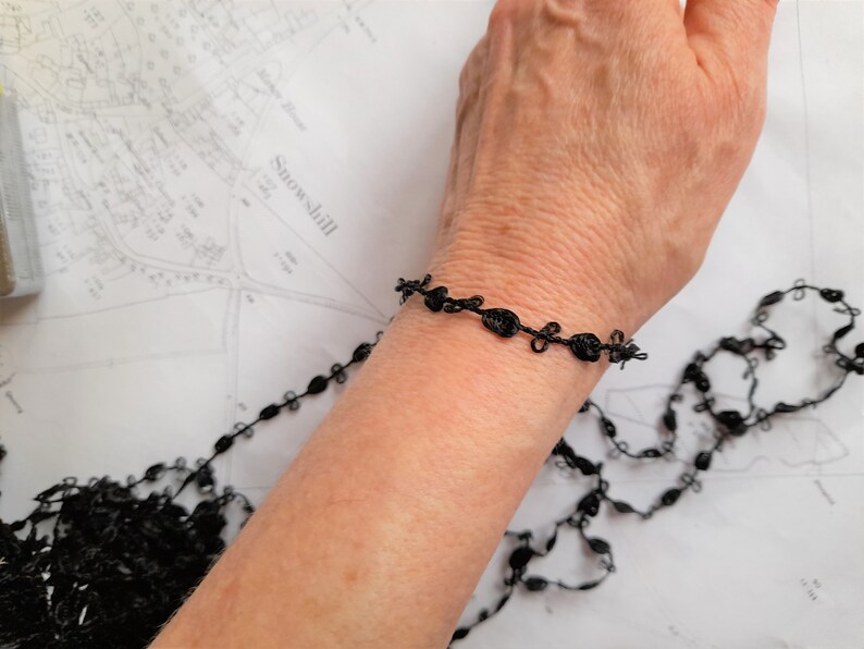 May include: A black lace bracelet with a delicate design. The bracelet is made of thin, black thread that is woven into a series of small, intricate knots.