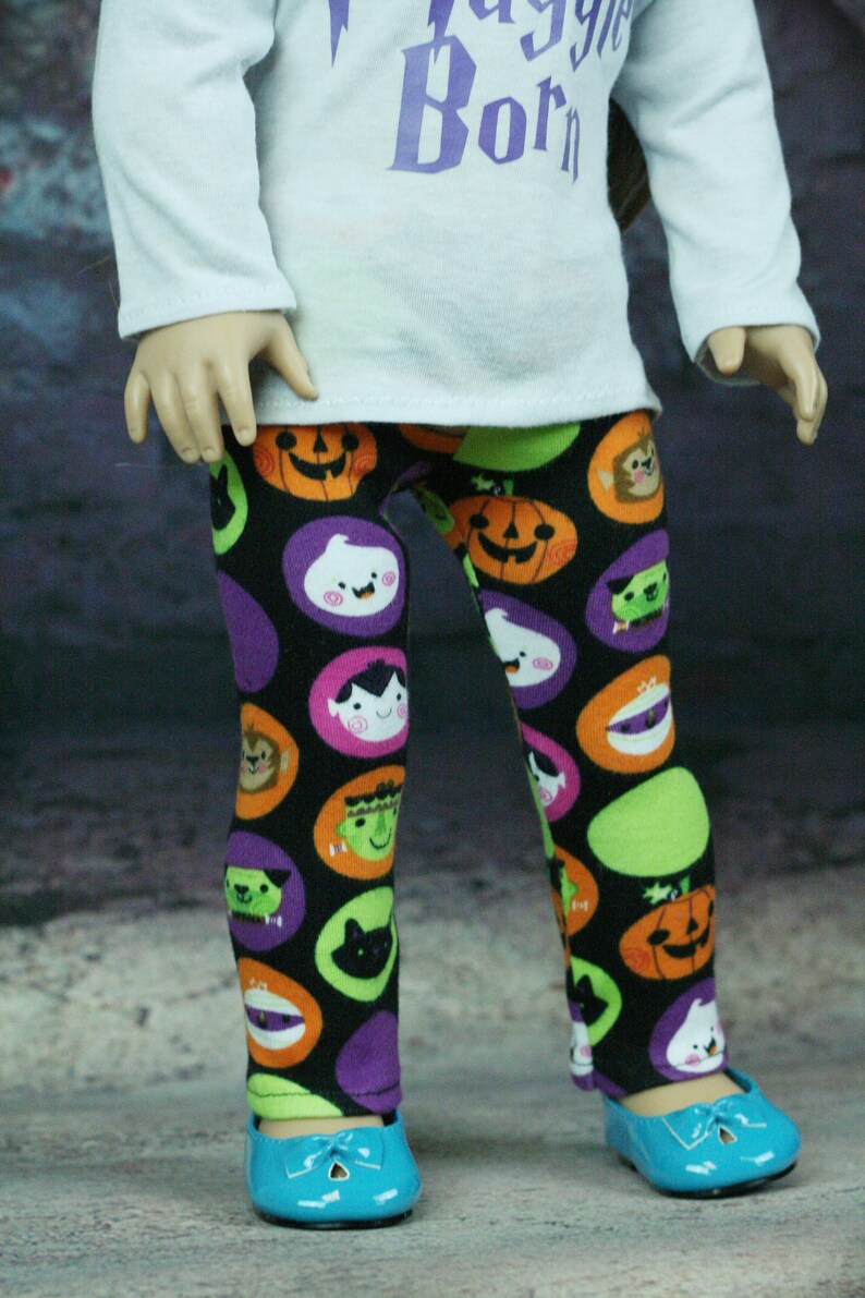 以下が含まれることがあります： ハロウィンをテーマにしたプリントが施された黒いレギンス。かぼちゃ、幽霊、その他の不気味なキャラクターが描かれています。レギンスは人形が着用しており、青い靴と組み合わせています。