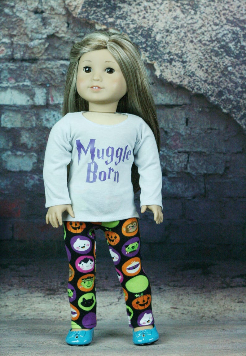 以下が含まれることがあります： 白い長袖シャツに「Muggle Born」という紫色の文字が書かれた人形。人形は、ハロウィンのキャラクターがカラフルな模様で描かれた黒いレギンスを履いています。人形は青い靴を履いています。