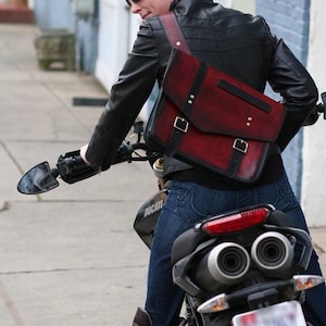 May include: A person wearing a black leather jacket and blue jeans is riding a motorcycle. They are carrying a burgundy leather messenger bag with black accents and buckles. The bag has a flap closure and a strap that goes over the shoulder. The motorcycle is black and has a silver exhaust pipe. The person is wearing sunglasses and has short, curly hair.