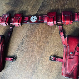 Puede incluir: Un cinturón de cuero rojo con múltiples bolsas y dos fundas para llevar armas de fuego. El cinturón está decorado con tachuelas metálicas y tiene una hebilla con acabado plateado.