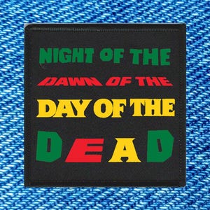 May include: A black square patch with the words "NIGHT OF THE DAWN OF THE DAY OF THE DEAD" in green, red, yellow, and green lettering. The patch is set against a blue denim background.