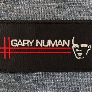 May include: Black and white embroidered patch with the text "GARY NUMAN" in white and two red lines. A white outline of a person's face is on the right side of the patch.