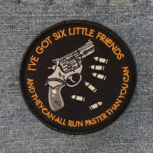 May include: A black, round embroidered patch featuring a silver revolver and six bullets. The patch has the text "I'VE GOT SIX LITTLE FRIENDS AND THEY CAN ALL RUN FASTER THAN YOU CAN" in orange lettering.