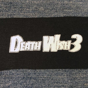 Può includere: Una toppa rettangolare nera con la scritta "Death Wish 3" in bianco, bordata da un colore più chiaro. La toppa è posta su uno sfondo di denim blu.