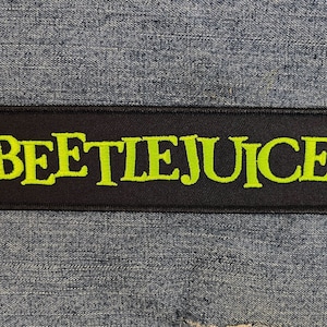 以下が含まれることがあります： 黒と緑の刺繍パッチで、スタイリッシュなフォントで「BEETLEJUICE」というテキストが書かれています。