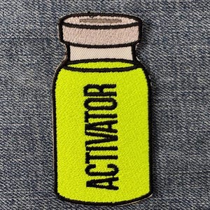 May include: Embroidered patch in the shape of a vial, with a light green interior and the word "ACTIVATOR" in black. The vial has a white and gray cap and a black outline. The patch is on a blue denim background.