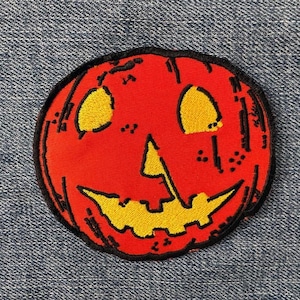May include: A round, orange pumpkin patch with a black outline and a smiling face. The eyes and mouth are yellow. The patch is on a blue denim background.