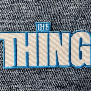 May include: Embroidered patch featuring the text "THE THING" in white block letters with a blue outline. The word "THE" is stacked above "THING". The patch is on a denim background.
