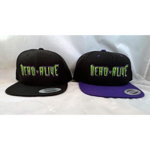 Puede incluir: Dos gorras de béisbol negras y verdes con el texto "Dead Alive" bordado en la parte delantera. Una gorra tiene una visera morada.