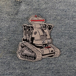 Puede incluir: Un robot gris y negro con una luz roja en la parte superior, sobre un fondo de mezclilla azul. El robot tiene un cuerpo de tanque con orugas y m&uacute;ltiples brazos.