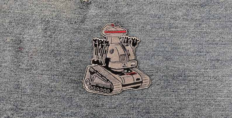 Puede incluir: Un robot gris y negro con una luz roja en la parte superior, sobre un fondo de mezclilla azul. El robot tiene un cuerpo de tanque con orugas y m&uacute;ltiples brazos.