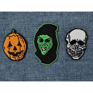 Könnte beinhalten: Drei Halloween-Stick-Patches. Ein orangefarbener Kürbis mit geschnitztem Gesicht, eine grüngesichtige Hexe mit schwarzer Kapuze und ein schwarz-weißer Totenkopf. Die Patches befinden sich auf einem blauen Jeanshintergrund.