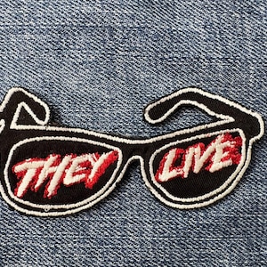 Puede incluir: Parche bordado con gafas de sol negras con la frase "THEY LIVE" en letras rojas y blancas. El parche tiene un borde blanco y está sobre un fondo de mezclilla azul.
