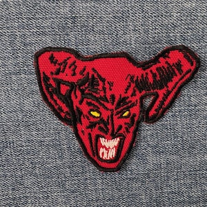 May include: A red embroidered patch featuring a devil's face with yellow eyes, sharp teeth, and horns. The patch is outlined in black and set against a blue denim background.