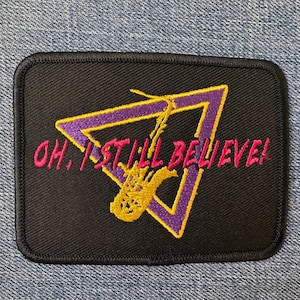 May include: A rectangular black embroidered patch featuring a purple triangle outline with a yellow saxophone graphic inside. The text "OH, I STILL BELIEVE!" is stitched in bright pink across the center of the design, which has a black border.