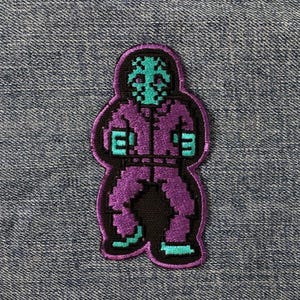 May include: A purple and black patch depicting a pixelated figure with a teal face and accents. The figure wears a purple jumpsuit and has teal hands and feet. This patch is likely an iron-on or sew-on accessory for clothing.