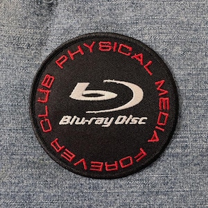 Pode incluir: Um remendo bordado preto e branco com o texto "Club Physical Media Forever" em vermelho e o logotipo do Blu-ray Disc em branco.