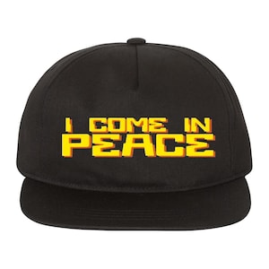 May include: Black baseball cap featuring the text "I COME IN PEACE" in yellow pixelated lettering. The cap has a flat peak and a rounded crown. The text is in a retro video game style.