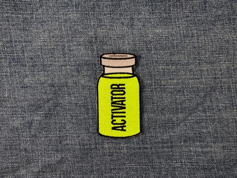 Peut inclure: &Eacute;cusson brod&eacute; repr&eacute;sentant un petit flacon jaune avec le mot "ACTIVATOR" en lettres majuscules noires. Le flacon a un bouchon marron clair et est soulign&eacute; en noir. L'&eacute;cusson est pos&eacute; sur un fond en denim.