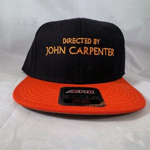 Puede incluir: Gorra de béisbol negra y naranja con el texto "DIRECTED BY JOHN CARPENTER" bordado en dorado. La gorra tiene una visera plana y una corona estructurada. La etiqueta dice "ONE SIZE FITS MOST".