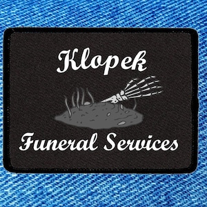 May include: Black rectangular patch with the text "Klopek Funeral Services" in white script. The patch features a gray illustration of a skeleton hand emerging from a mound of dirt. The background is a blue denim texture.