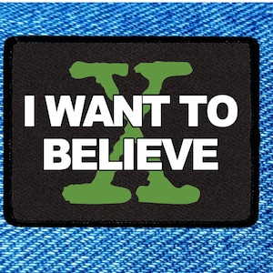 May include: A black rectangular patch with the text "I WANT TO BELIEVE" in white letters and a green "X" symbol. The patch is set against a blue denim background.