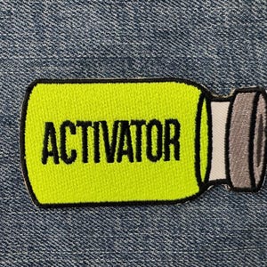 Peut inclure: &Eacute;cusson brod&eacute; en forme de flacon, avec un centre jaune vif et une bordure noire. Le mot "ACTIVATOR" est imprim&eacute; en lettres noires en gras. Le bouchon du flacon est gris. L'&eacute;cusson est pos&eacute; sur un fond en denim bleu.