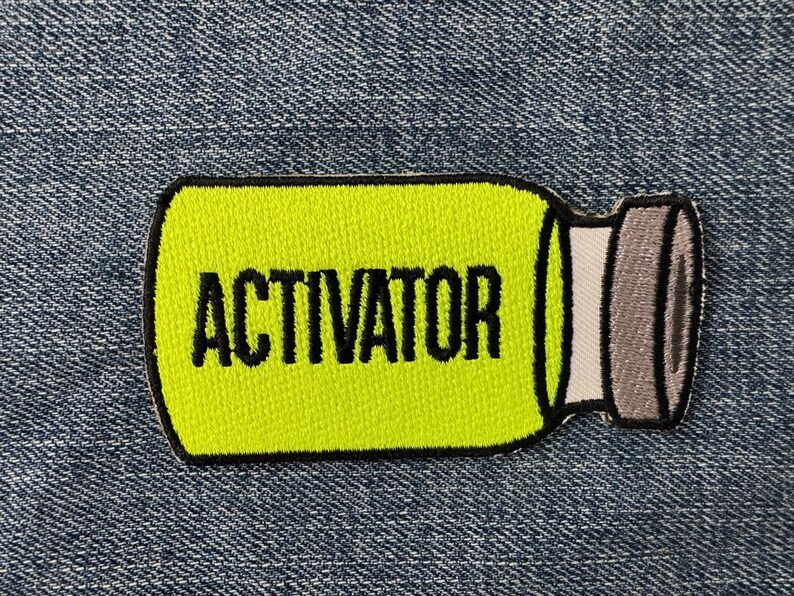 Peut inclure: &Eacute;cusson brod&eacute; en forme de flacon, avec un centre jaune vif et une bordure noire. Le mot "ACTIVATOR" est imprim&eacute; en lettres noires en gras. Le bouchon du flacon est gris. L'&eacute;cusson est pos&eacute; sur un fond en denim bleu.