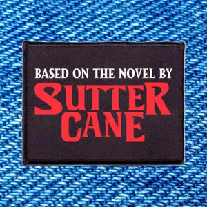 May include: A rectangular black patch with white text that reads BASED ON THE NOVEL BY and red text that reads SUTTER CANE. The patch has a stitched border and is displayed against a textured blue denim fabric background.