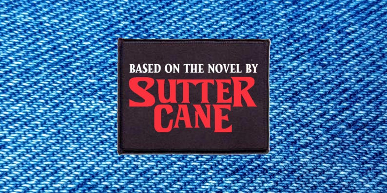 以下が含まれることがあります： BASED ON THE NOVEL BYという白い文字とSUTTER CANEという赤い文字が書かれた長方形の黒いワッペン。ワッペンにはステッチの縁取りがあり、質感のある青いデニム生地を背景に配置されています。