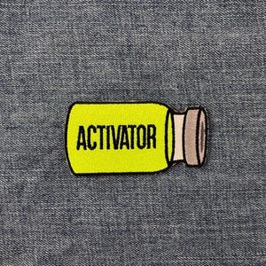 Peut inclure: Un &eacute;cusson jaune en forme de bouteille avec le mot "ACTIVATOR" en noir. La bouteille a un bouchon gris et blanc. L'&eacute;cusson est pos&eacute; sur un fond en denim bleu.