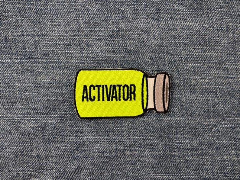 Peut inclure: Un &eacute;cusson jaune en forme de bouteille avec le mot "ACTIVATOR" en noir. La bouteille a un bouchon gris et blanc. L'&eacute;cusson est pos&eacute; sur un fond en denim bleu.