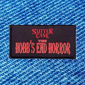 May include: A rectangular black patch with red embroidered text that reads SUTTER CANE THE HOBB'S END HORROR. The patch features a bold, stylised font for the title and a smaller font for the author name, all centred on a dark fabric background.