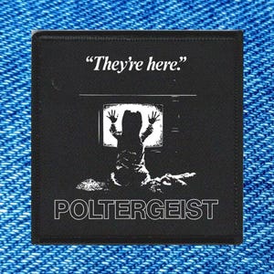 May include: Black square patch with a graphic from the film Poltergeist. The patch features the text "They're here." above an image of a child reaching out from a television screen. The word "POLTERGEIST" is printed below the image.