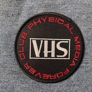Pode incluir: Um remendo circular preto e vermelho com o texto "VHS Physical Media Club Forever" em letras vermelhas ao redor da borda. O centro do remendo tem um retângulo branco com o texto "VHS" em letras brancas.