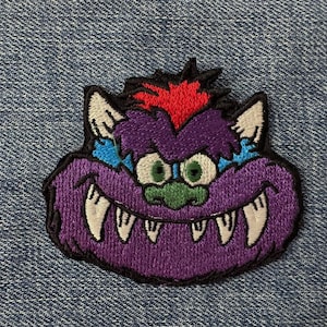 Może przedstawiać: Haftowana naszywka przedstawiająca twarz potwora z kreskówki. Potwór ma fioletowe futro, białe kły, zielone oczy i czerwony irokez. Naszywka znajduje się na niebieskim tle z dżinsu.