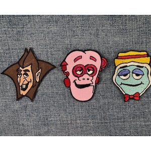 May include: Three embroidered patches featuring cartoon characters. The first is a brown and tan vampire with a star-shaped background. The second is a pink monkey-like face. The third is a blue face with a red bow tie and a yellow and red hat.