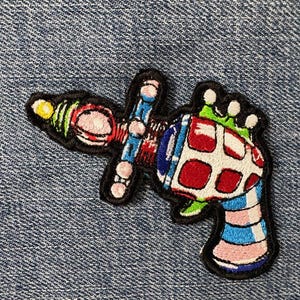 May include: Embroidered patch depicting a colourful ray gun design. The patch has a black border and features red, white, blue, pink, green, and yellow details. The ray gun has a whimsical, cartoonish style.