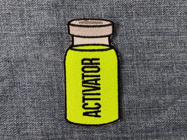 Peut inclure: &Eacute;cusson brod&eacute; en forme de flacon, avec un int&eacute;rieur vert clair et le mot "ACTIVATOR" en noir. Le flacon a un bouchon blanc et gris et un contour noir. L'&eacute;cusson est sur un fond en denim bleu.