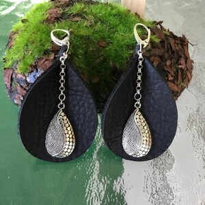 May include: A pair of black leather earrings with silver teardrop charms. The charms have a textured design and are attached to the earrings with a silver chain.